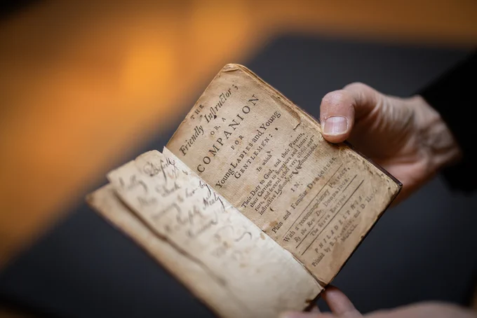 A person standing just off camera holds out an old, faded book to its title page. Title is, "A Friendly Instructor; Or A Companion for Young Ladies and Young Gentlemen."