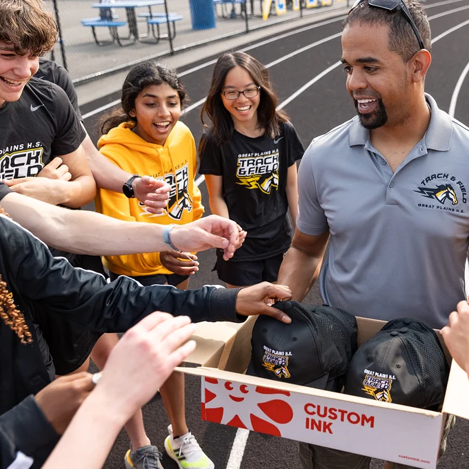 Adult holding a Custom Ink box filled with custom hats. Students surround the adult to pick up a hat.