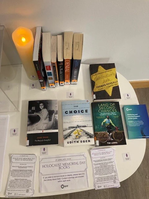 A photo of the BPP Holborn Library Space of Reflection-a series of books chosen to commemorate Holocaust Memorial Day are shown face up on a white table that also displays guidelines and information on the Space of Reflection, HMD logo stickers, and a white commemorative non-flammable electronic candle (switched on to emit a candle-like glow).