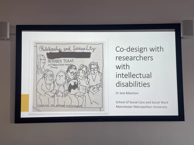 Photo of the power point on the big screen it is the introductory slide and says foreign with researchers with ID with my name and social care and social work in MMU and an image which is a hand drawn image of the research team@