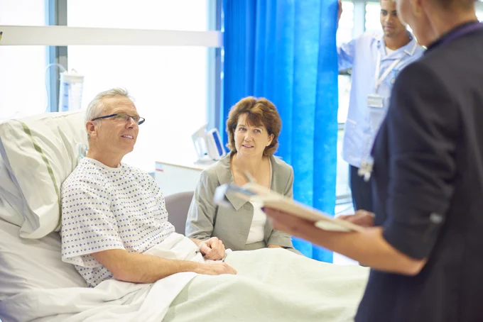 A man is sitting in a hospital bed with his wife beside him while a consultant speaks to him about his procedure