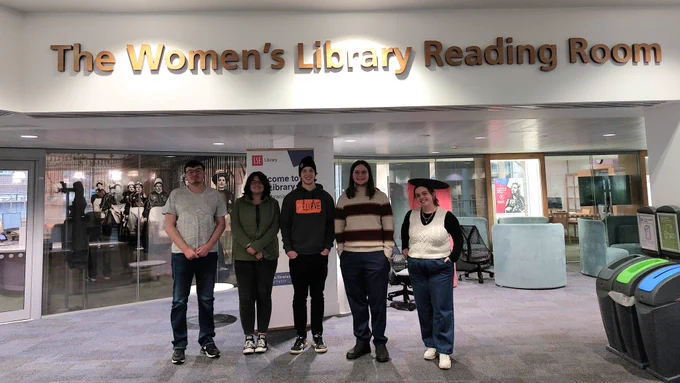 Five people stood for a photograph outside The Women's Library Reading Room on the 4th floor of LSE Library. Above them is the Women's Library signage and behind them is a photo of suffragettes marching.