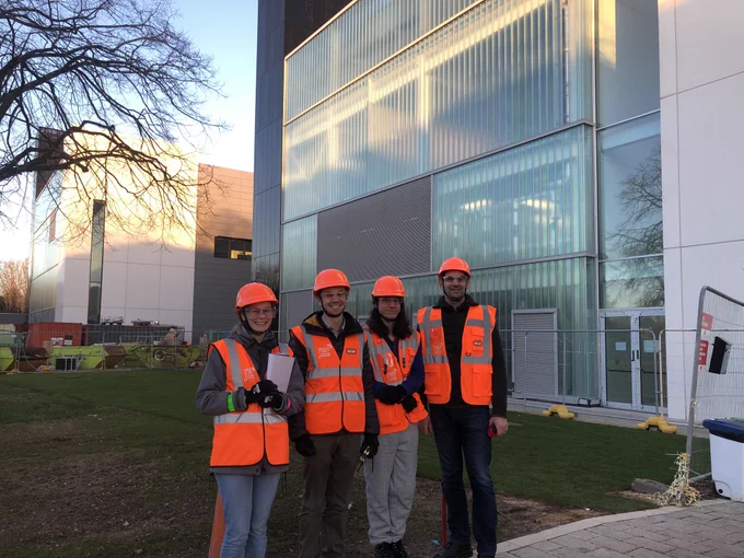 Four people in high vis orange jackets and hard hats are standing outside a glass building in the sunset. There are metal barriers and skips in the background to show it’s still a building site but from the outside at least it looks complete!