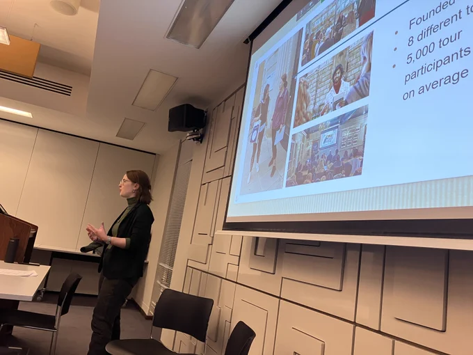 Olivia stands in front of a desk, talking to an unseen group. Behind her a slide shows pictures of Uncomfortable Oxford.