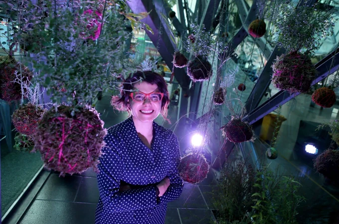 Dr Tanja Beer, a smiling white woman with brown hair to her ears, red glasses, and a blue shirt with white polka dots. She stands in a dimly lit space, with plants rooted in spheres of what looks like soil hanging from the ceiling all around her.