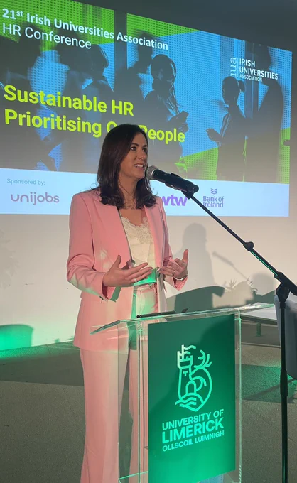 Laura Ryan standing in a pink suit speaking at a green podium