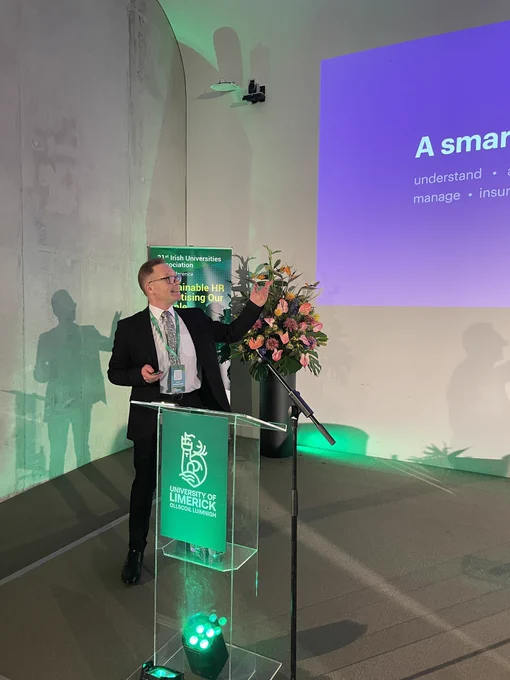 Professor Michael Morley speaking while pointing to a large digital screen. He is standing beside a podium.