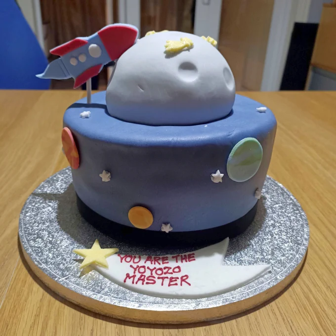A decorative cake shaped like outer spec with a protruding planet and stars and a rocket ship. below a shooting star appears with the message "YOU ARE THE YOYOZO MASTER"