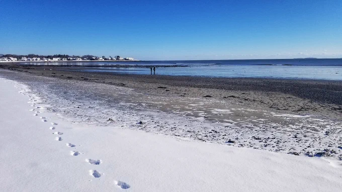 Sandy beach with snow in the foreground and water and houses in the background.