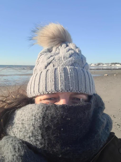 Headshot of a person standing on a sandy beach wearing a hat and scarf.