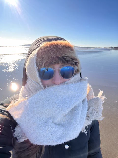 Headshot of a person standing on a sandy beach wearing a hat, scarf and sunglasses.