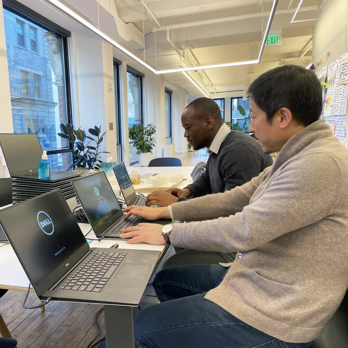 two men working at a table with three open laptops and a pile of laptops behind