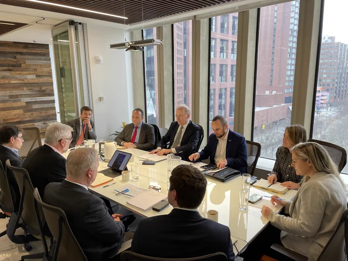 A group of people sit around a conference table. Outside the windows behind them are buildings. 