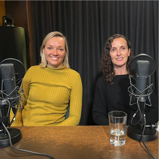 A photo of Susanne Gedamke and Stefanie Brown sitting behind two microphones in a recording studio.