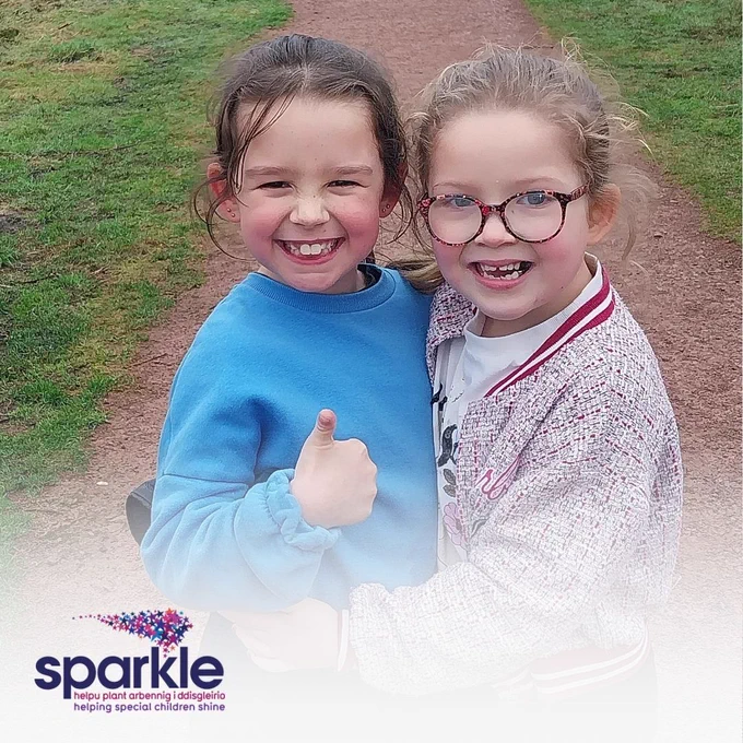 Two young girls stand side by side. The girl on the left has brown hair tied back and wears a blue jumper - she grins and gives a thumbs up to the camera whilst other girl hugs her, the girl on the right wears a multicoloured jacket and black/pink glasses. 