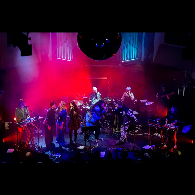 Eight musicians on stage playing instruments and singing. The stage is backlit by red and blue lighting.