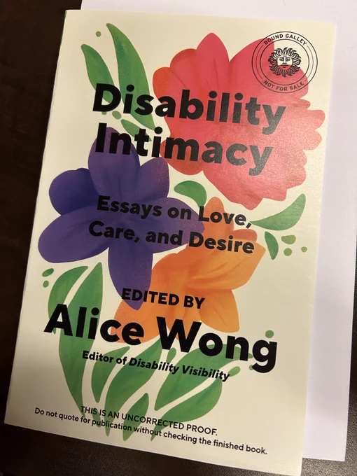 a paperback galley of Disability Intimacy: Essays on Love, Care, and Desire edited by Alice Wong on a wood desk background. cover is cream-colored with green leaves and large red, purple, and orange watercolor flowers