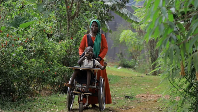 A young woman in a red dress and green hijab is walking down a jungle path pushing her young brother in a supportive wheelchair.