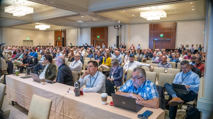 A crowd of people taking notes on their computers or notebooks while actively listening to a 5x5 panel in a large conference room