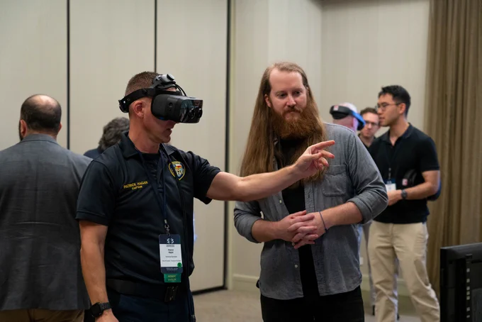  
One man testing a VR head said while another observes. Others are testing VR goggles behind.
