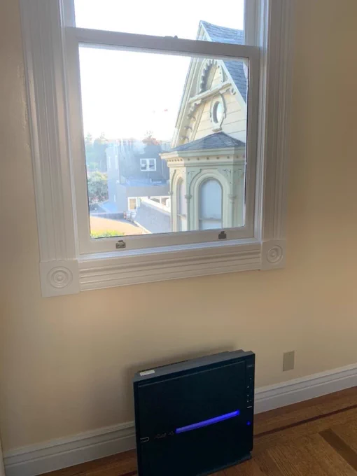 A MinusA2 air purifier is positioned on the floor of a room with hardwood floors and a large window that frames a view of a historic-looking building with intricate architecture.