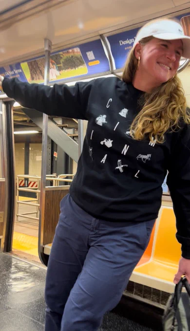 Phoebe Judge wearing a black Criminal sweatshirt and white visor and smiling widely. She leans toward the right side of the image, holding on to a pole in a subway car to support herself.