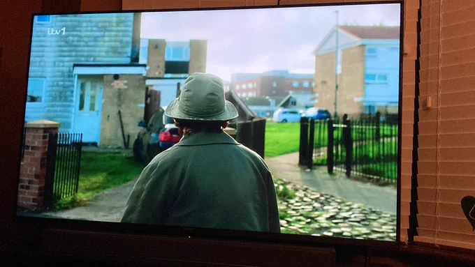 A still from TV detective programme Vera showing a street scene on a housing estates. The roof of the hospital can be seen in a gap in the houses. Vera stands with her back to the camera in her green Mac and hat.