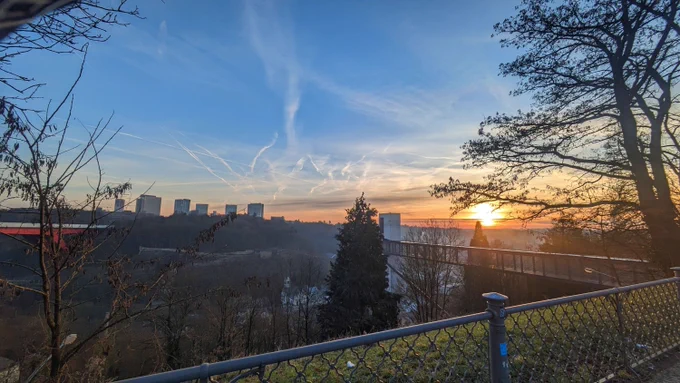 View from the top of a hill, a sun is rising in the background with orange yello colors that reflect on the small clouds and contrails. Shadows of trees frame the view. Part of the Luxembourg red bridge can be seen on the right, with some silhouettes from high buildings in the Kirchberg. The light is gold yellow, giving a comfy feeling to the whole scene.