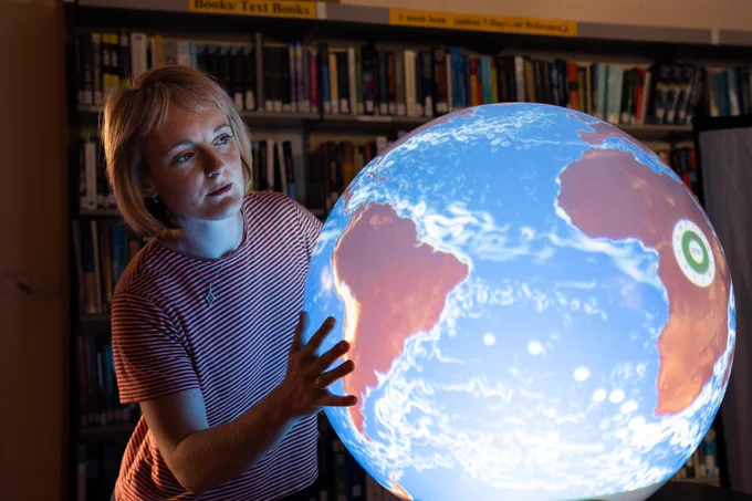 Person with shoulder-length hair and a striped shirt looks closely at a large illuminated globe, in a dark room. They touch the globe with both hands.