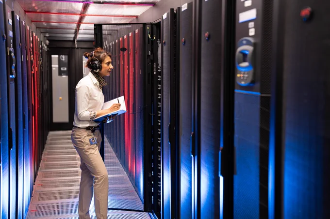 Person holds a pen and paper pad, wearing ear defenders in a large aisle full of computer equipment.