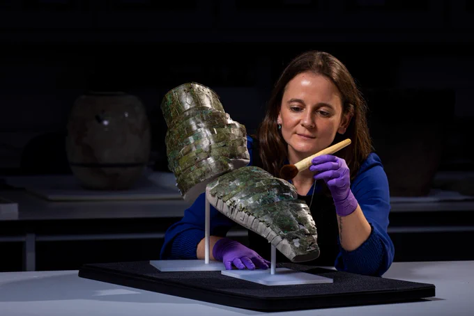 Conservator Bethan Bryan with a Roman arm guard mounted to look like it may have once looked on a soldier, covering the upper and lower part of the sword arm. She uses a large brush to clean the greenish brass.