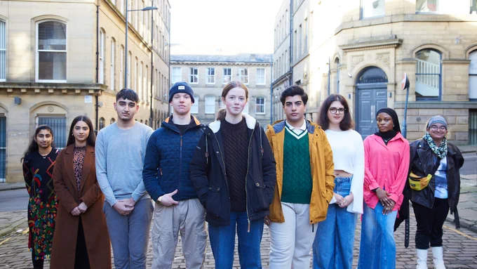 ID: Image shows diverse group of young people stood on Bradford's cobbled streets in a line. 