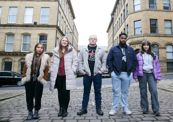 ID: Image shows diverse group of young people stood on Bradford's cobbled streets in a line. 