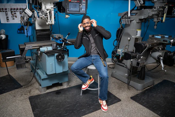Baran Mensah smiles while putting on lab safety glasses. He sits on a stool surrounded by large machinery in a lab.