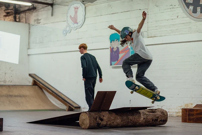a young person is jumping over a skateboard ramp (on their skateboard). They are inside and the ramp is made of a log and metal sheets