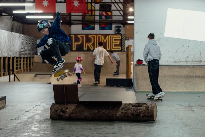 another young person is jumping over a skateboard ramp (on their skateboard). They are inside and the ramp is made of a log and metal sheets. Behind them are more young people skateboarding. 