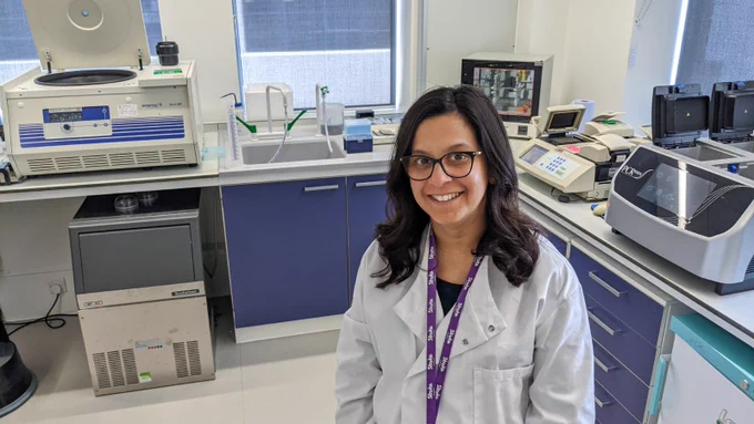A researcher in a white coat standing in her lab. She's smiling at the camera and is wearing a Stroke Association lanyard. 
