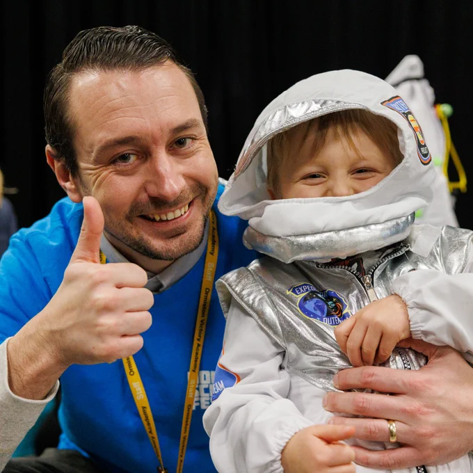 Small child dressed as astronaut with a grown-up giving the thumbs-up. 