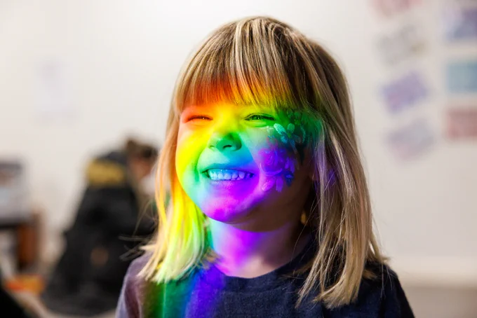 Little girl grinning, with a rainbow light cast onto her face. 