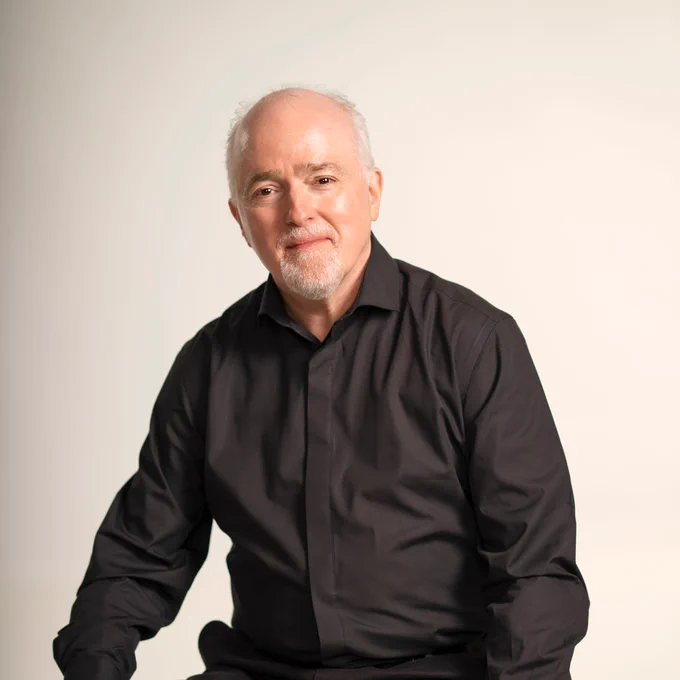 Paul Murphy is sitting on a aged wooden stool, smiling for a headshot. Paul is facing forwards, looking at the camera. Paul has grey/white short hair and a trimmed goatee. Paul is wearing a black formal shirt, black trousers and a gold watch sits on the left wrist. Credit: Johan Persson.