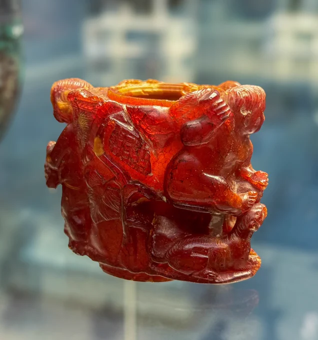 Rear of the red and orangish amber perfume pot, with light streaming through the stone. Here, we can see a winged cupid on either side, and a panther lying below the Cupid on the right. On the front are grape vines and a kantharos (traditional Greek wine cup of the god Dionysus-Bacchus). The top has a circular opening, and the stone has been carved out to store perfume. It would have had a plug or stopper once, now lost.

This perfume pot is from Aquileia, which was the center of the amber trade in the Roman world. Workshops in the city created beautiful objects like this from raw amber, which was then sold throughout the empire. 

Photo: my own