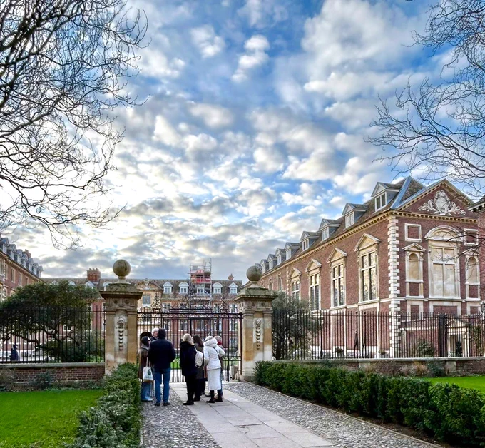 A group of tour attendeees stand outside the red brick buildings of St. Catz College in Cambridge listening to an Uncomfortable Cambridge tour guide. It is a nice day with blue skys and beautiful clouds.