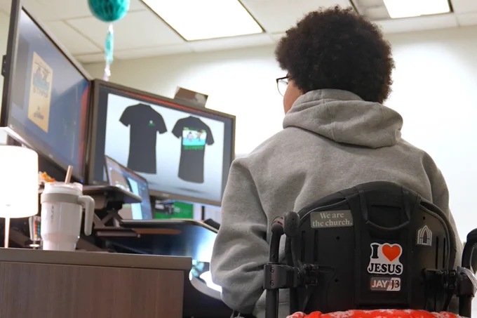 Jayden sits at a desk to work with the Stars designer to design his tshirt, which is clearly shown on the screens of the computers