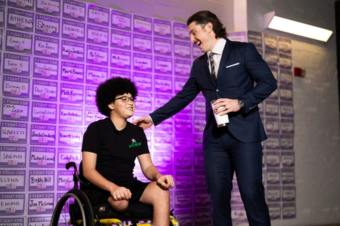 Matt Duchene and Jayden laugh when talking on hockey fights cancer night. Jayden is in his chair and Matt is standing in his suit smiling and laughing, reaching for a hug
