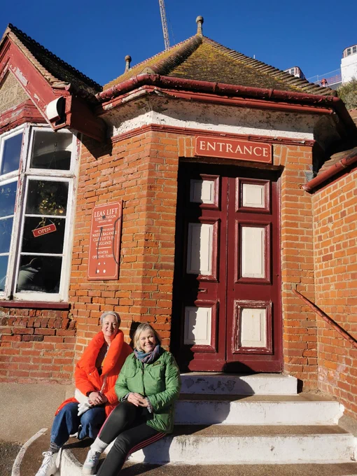 Two women - Amanda Beckett and Charlotte Chapman - sat on the steps in front of the entrance to the Leas Lift in Folkestone. There is blue sky in the background.