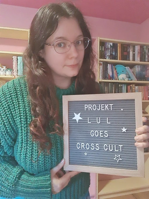 Selfie von mir, braunes, lockiges Haar, Brille, grüner Pullover, mit Schild das sich liest: Projekt Lul goes Cross Cult