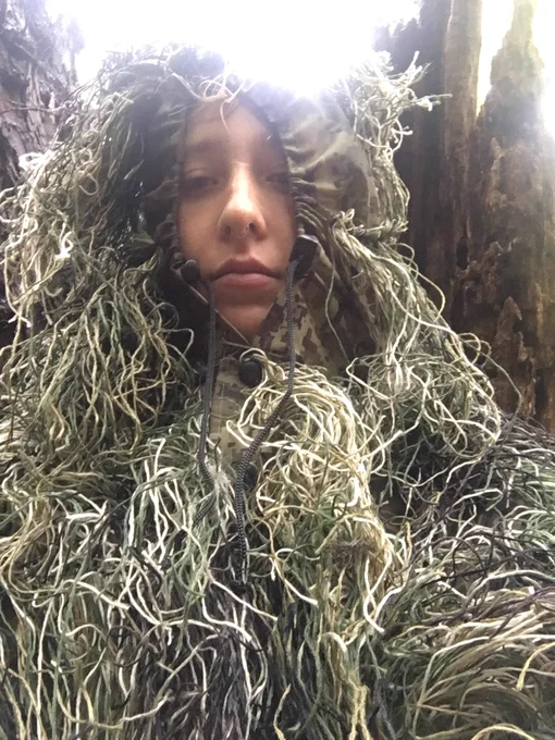selfie of jess, a white woman in an oversized ghillie suit, in the forest