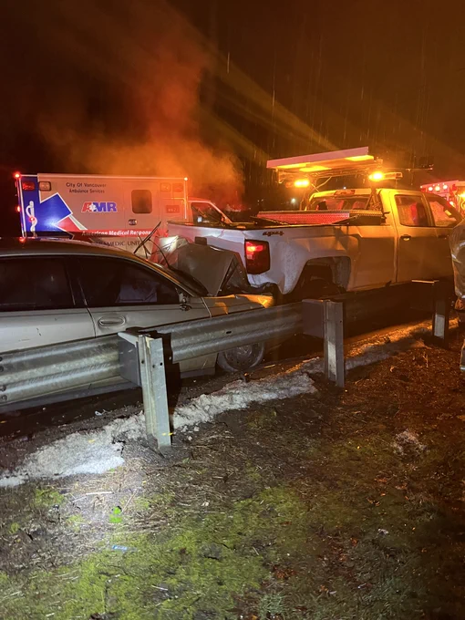 A car crashed into the back of a white pickup work truck at night on a highway with emergency responders around.