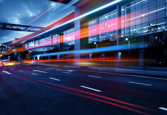 A long shot of a road with a glass building next to it, with beams of light suggesting movement across the road.