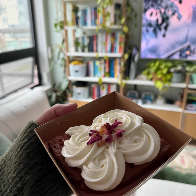 A photo of the celebratory delicious rose milk cake with white frosting and rose petals on top held up against the backdrop of Fatima's living room - lots of books, plants, and a soft winter landscape painting on the tv. 